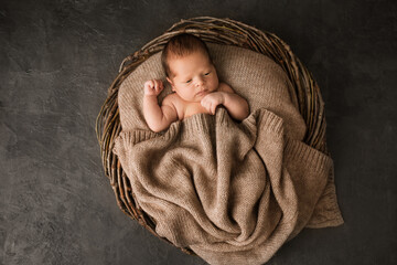 A newborn baby covered with a warm knitted blanket in a wicker basket - obrazy, fototapety, plakaty