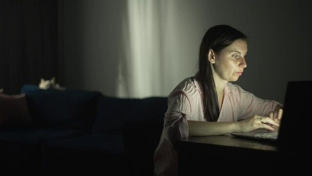 The girl works at the computer in the evening at home. A woman is typing on a laptop and a dog is resting on the sofa. Remote work. High quality 4k footage