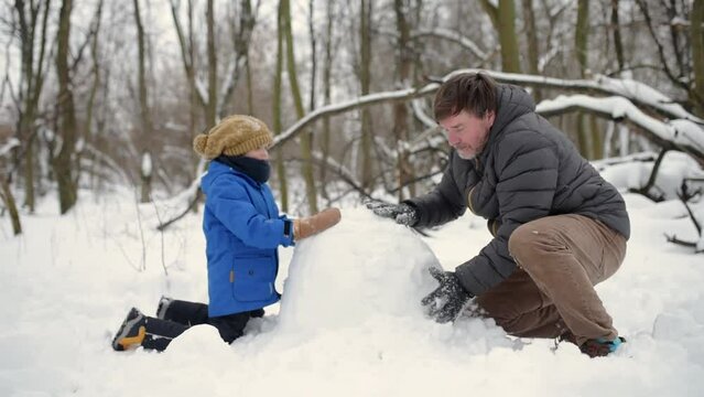 Little boy with his father building snowman in snowy park. Dad and son spend quality time together. Active outdoors leisure with family with children in winter. Kid during stroll in a snowy winter par