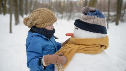 Little boy building snowman in snowy park. Child puts hat and scarf on snowman . Active outdoors leisure with family with children in winter. Kid during stroll in a snowy winter park - Powered by Adobe