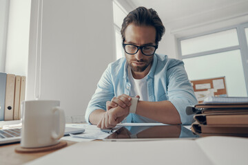 Fototapeta premium Handsome young man looking at his smart watch while sitting at his working place in office