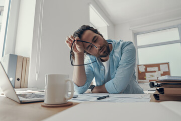 Tired young man keeping eyes closed while sitting at his working place in office