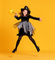 Happy girl in  witch costume with pumpkin basket jumping and celebrates Halloween on yellow...