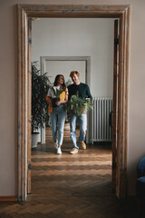 Happy young couple holding houseplants while walking by their new apartment together - obrazy, fototapety, plakaty