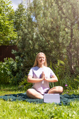 A young blonde girl is sitting on the grass in the park and doing yoga online