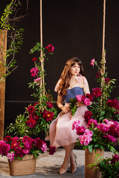an attractive young woman in a dress sits on a swing in peony flowers. 