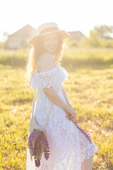 Beautiful young girl in a white dress, straw hat, with picnic and bouquet of purple wild flowers on a meadow. Summertime, golden hour, sunset