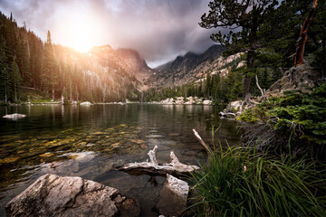 Mountain lake at sunset with granite rock and trees along the shoreline. Orange sunlight is shining down onto the lake from the top of a mountain peak. 