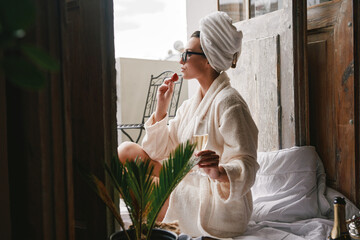 Beautiful young woman in bathrobe enjoying strawberry and champagne while relaxing on the balcony