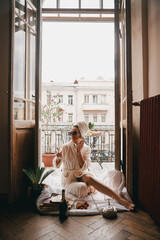 Happy young woman in bathrobe using phone and enjoying champagne while sitting on the balcony