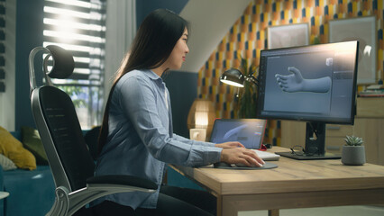 Asian woman sitting on chair at the table at home office and making 3D prototype of human hand in professional program for 3D modelling, using modern computer and digital tablet.