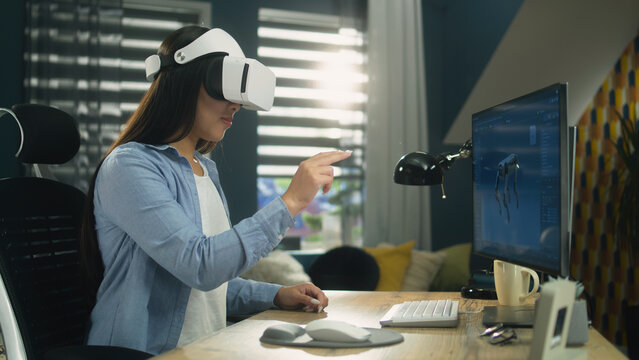 Asian woman sitting at the table at home office and using VR headset to work on 3D modeling project on personal computer. Virtual reality. Metaverse