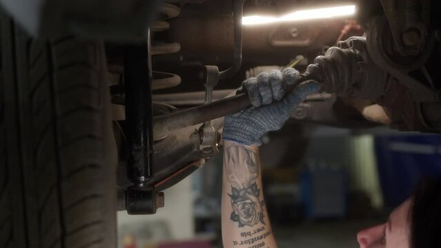 Close up hand mechanic in car service checks suspension of car that is raised on lift in repair zone shop. Vehicle maintenance for further admission to roads. Tatoo sign Acts are stronger than words