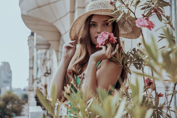 Beautiful young woman in elegant hat smelling flowers while standing on the balcony