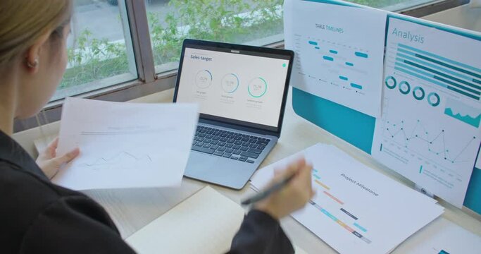 Business woman entrepreneur making data analysis on success plan strategy. Skilled female manage project study paper reports analyze stats infographic on computer screen and paperwork. Rear back view.