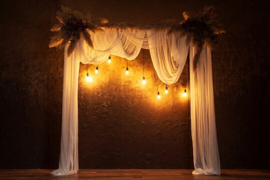 arch with white fabric against a brown wall with flowers. wedding classic arch for a wedding photo shoot. wooden arch with dry flowers and incandescent lamps in a photo studio