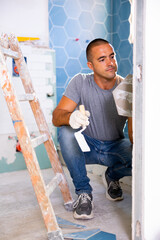 Portrait of concentrated young man sitting squat and making renovation repair at bathroom. Tiler...
