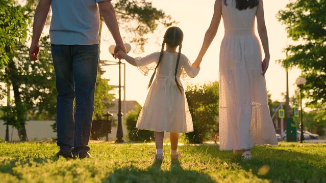 Family, child walking hand in hand in city park on green grass in sun. Slow motion. Active Family, mom daughter, dad play together outdoors. Happy childhood, future of child. Family walk concept, day
