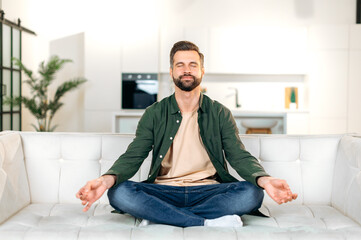 Satisfied calm happy caucasian bearded man, in a casual clothes, sits on a sofa in lotus position...