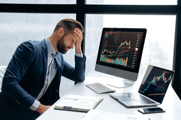 Upset stressed worried caucasian male stock investor, broker, financial expert, sit at work desk in front of computer with charts, nervous of fall cryptocurrency coin, closed eyes, disappointment