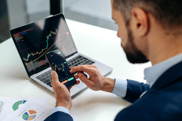 Close-up of laptop and smart phone screen with stock diagrams.Caucasian crypto trader investor using cellphone and laptop for cryptocurrency financial market analysis, buying or selling cryptocurrency