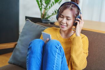 Fototapeta premium Portrait of a smiling Asian woman wearing a pair of headphones and using her phone and listening to music while sitting on the sofa