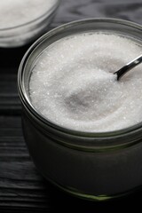 Granulated sugar and spoon in glass jar on black wooden table, closeup