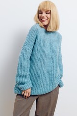  attractive woman in a beautiful light blue, long, knitted sweater stands on a light background in a relaxed pose and smiles pleasantly, turning her face to the side with her eyes closed
