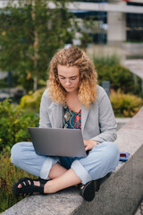 Caucasian curly woman university student sitting outdoors working on laptop computer, reading. Technology, education and remote working concept. Online