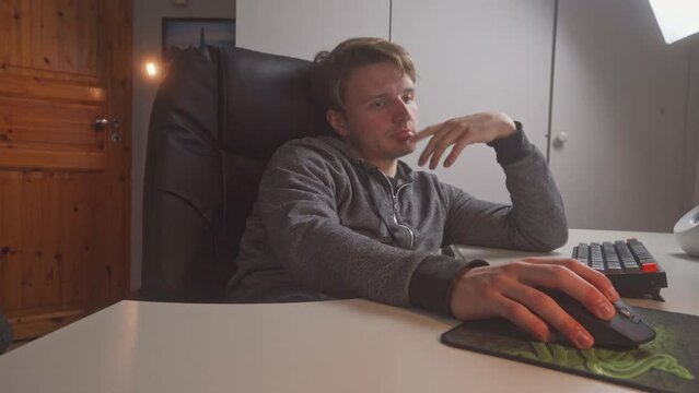 A young man is using his fingers to flip his lips as he sits in front of his home computer.  He is both bored and engaged in deep thought on how to build his social media presence