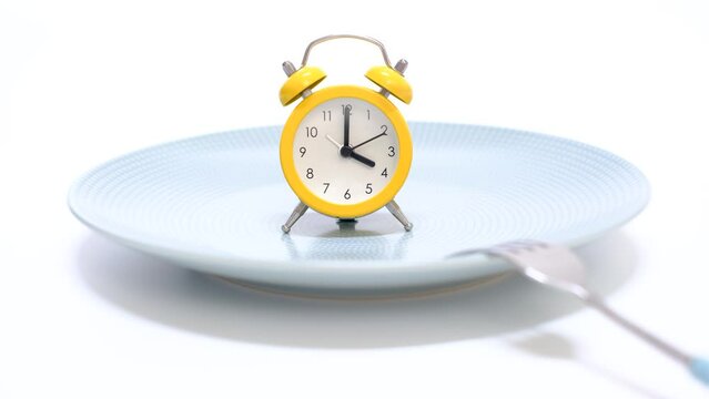 Empty plate with alarm clock and fork on white background, intermittent fasting concept.