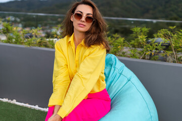 Stylish fit fashion women in bright pink wide leg pants and yellow shirt holding bag trendy sunglasses posing at rooftop terrace tropical view outdoor holding mobile phone