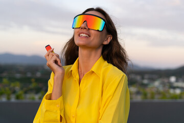 Stylish fit fashion women in bright yellow shirt trendy shield visor rainbow sunglasses posing at rooftop terrace tropical view sunset  smoking vape pod enjoying puffs