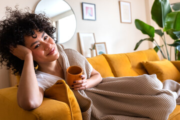 Happy African American woman relaxing at home lying on couch holding cup of warm coffee looking at...