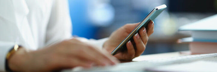 A woman works at a computer and holds a smartphone