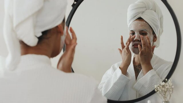 Young Black woman wearing robe and towel on head after taking morning shower standing in front of mirror fixing moisturizing sheet mask on her face