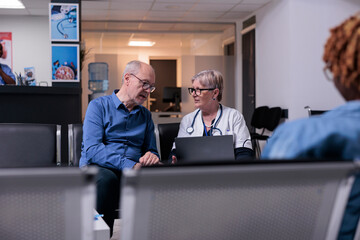 Physician using laptop at checkup visit with old man, sitting in waiting room lobby. General practitioner and patient talking about disease diagnosis and healthcare, consulting adult at health center.