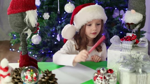 A little girl in a red Santa's Hat writes a letter to Santa Claus with her wishes under the Christmas tree. Christmas and New Year atmosphere
