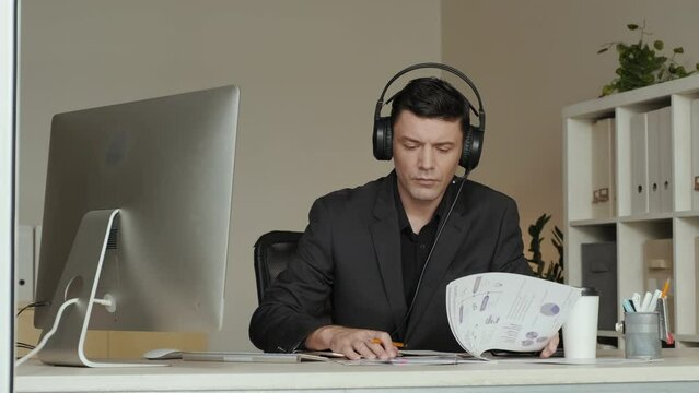 Medium shot of Caucasian businessman wearing headset with microphone working with business documents while sitting in front of computer in office