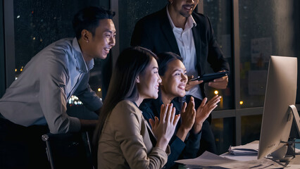 Group of Asian businesspeople looking at computer screen and celebrating success together in the office after working late at night.