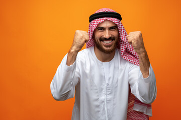 Successful smiling young Arab man celebrating victory on orange background