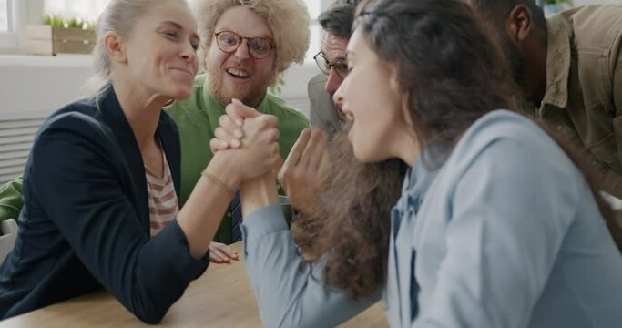 Businesswomen are competing in arm wrestling game in office enjoying break laughing having fun together while male colleagues are cheering.