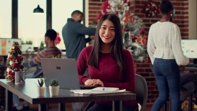 Portrait of female worker using laptop in festive office filled with winter decorations and ornaments. Woman working on startup during christmas holiday festivity in space with xmas tree.
