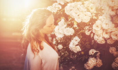 a beautiful woman smells a rosebush at sunset.