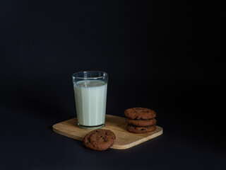 Cookies made from cocoa and with pieces of chocolate inside and glass of milk on a wooden board on a black background. Breakfast eating. Copy space