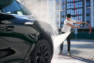 Guy in cap cleaning his black car using high pressure water in service. Washing of auto with...