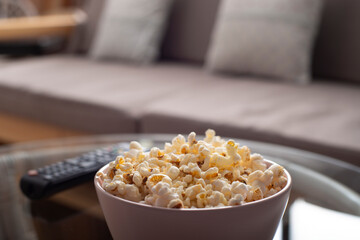 Bowl with popcorn and tv remote control on glass table near sofa