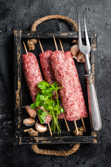 Fresh raw beef meat kebabs sausages on skewers in wooden tray. Black background. Top view