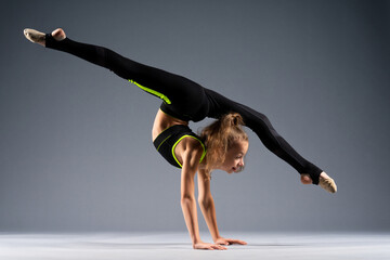 Flexible little girl doing acrobatic stunts while standing on her hands. The gymnast is dressed in a black tracksuit for stretching. Isolated on a gray background.