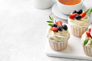 Vanilla cupcakes with buttercream and fresh berries, green leaves on grey background with tea. Delicious homemade dessert. Festive bakery.
